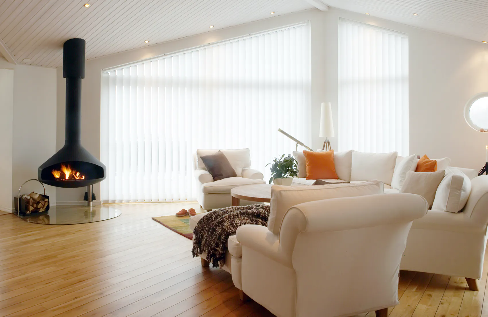 Lamellgardiner dekker store skråstilte vinduer og slipper inn mykt, diffust dagslys. I en lys, moderne stue med hvite sofaer, tregulv, oransje puter og hengende svart peis.