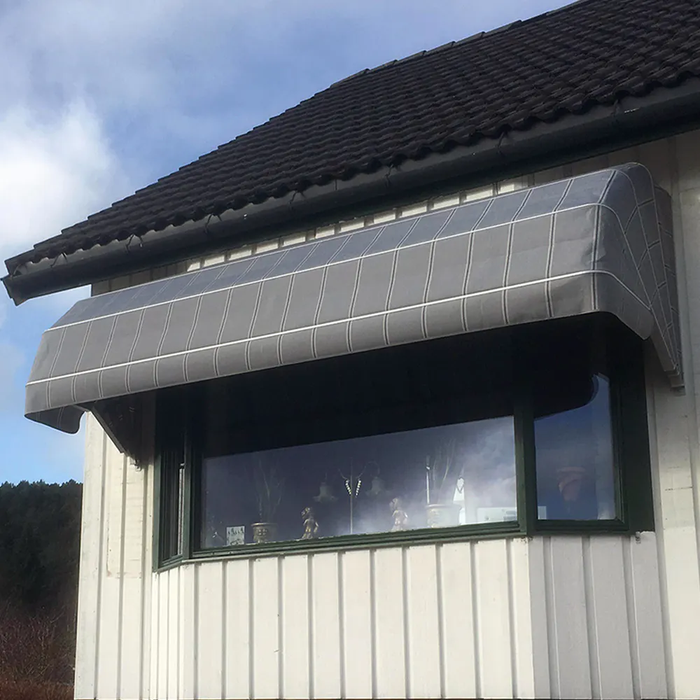 Gråstripet markise dekker og skygger et buet karnappvindu, montert under mørkt tak på hvit panelfasade, og beskytter mot sol og vær; bakgrunn av blå-hvit himmel og trær.