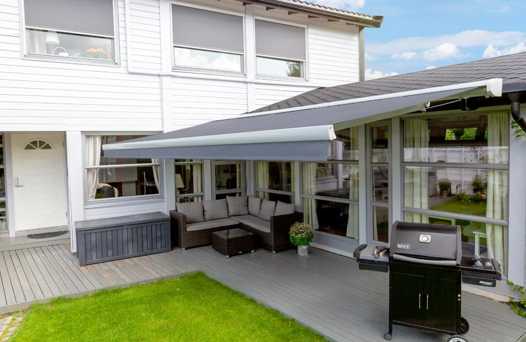 Markise (terrassemarkise) skyggelgger over uteplass; den dekker hjørnesofaen på et grått tredekke ved en hvit toetasjes bolig, med grønn plen og en Weber-grill i forgrunnen. Tekst: WEBER.