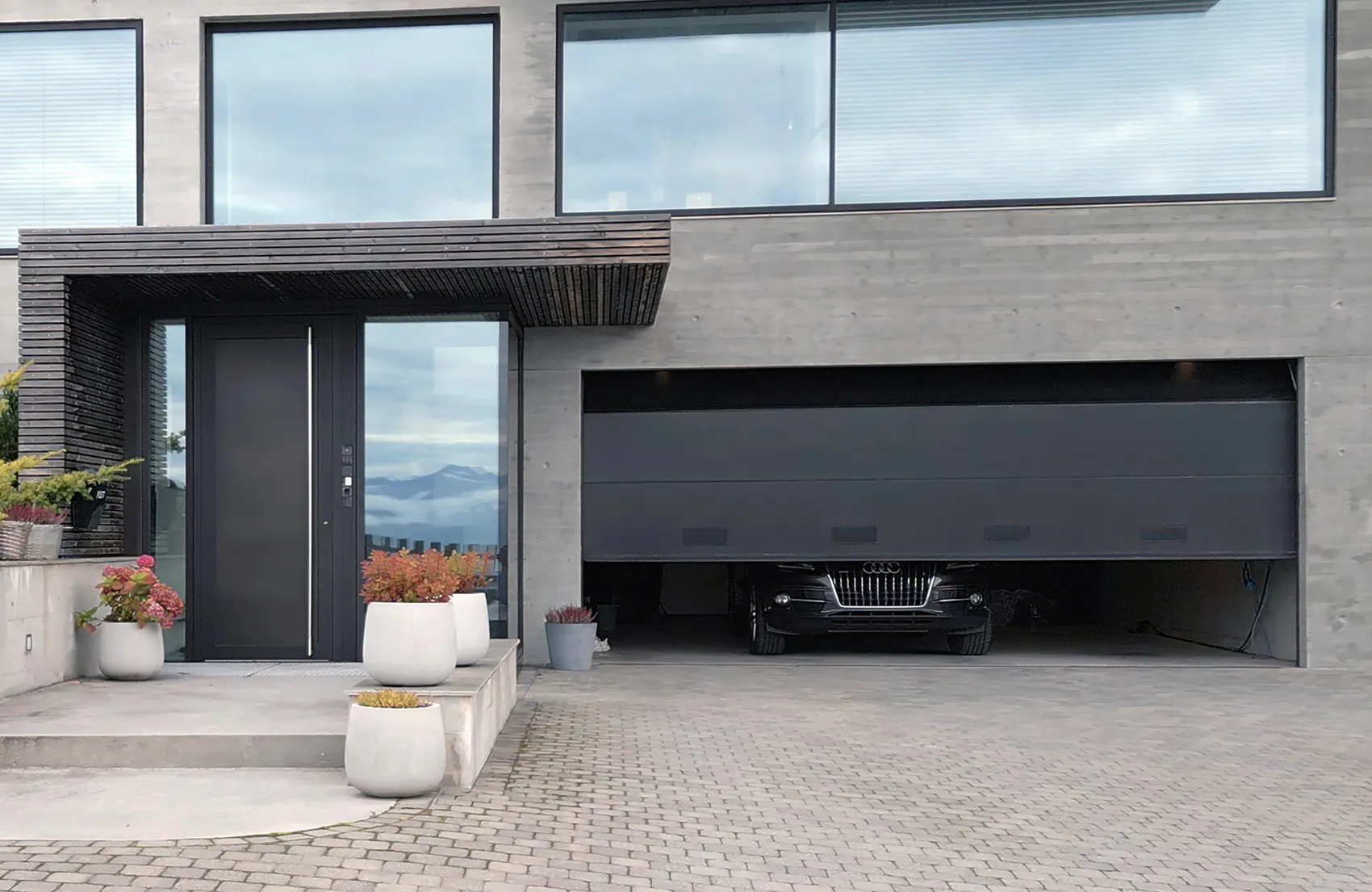 Garasjeport delvis åpnet, avdekker fronten på en svart bil; moderne grå betongfasade med store vinduer, inngangsdør og potteplanter på belegningsstein foran huset.