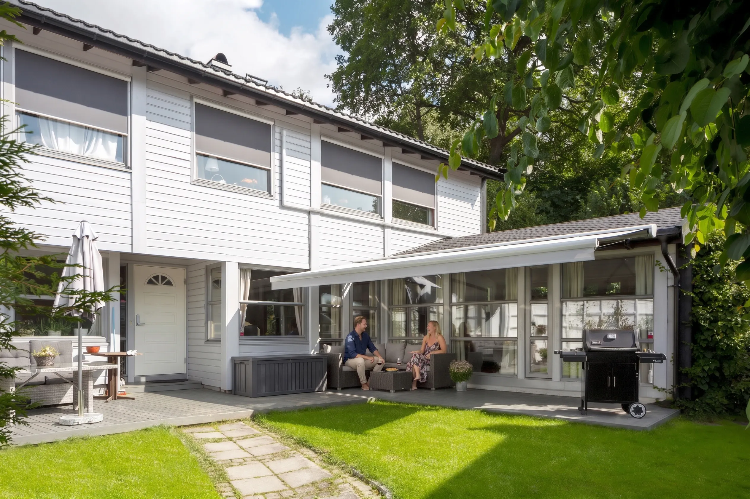 Utvendig markise er trukket ut og gir skygge over en sittegruppe. To personer sitter på terrassen ved en hvit to-etasjes bolig med glassveranda, gressplen og grill.