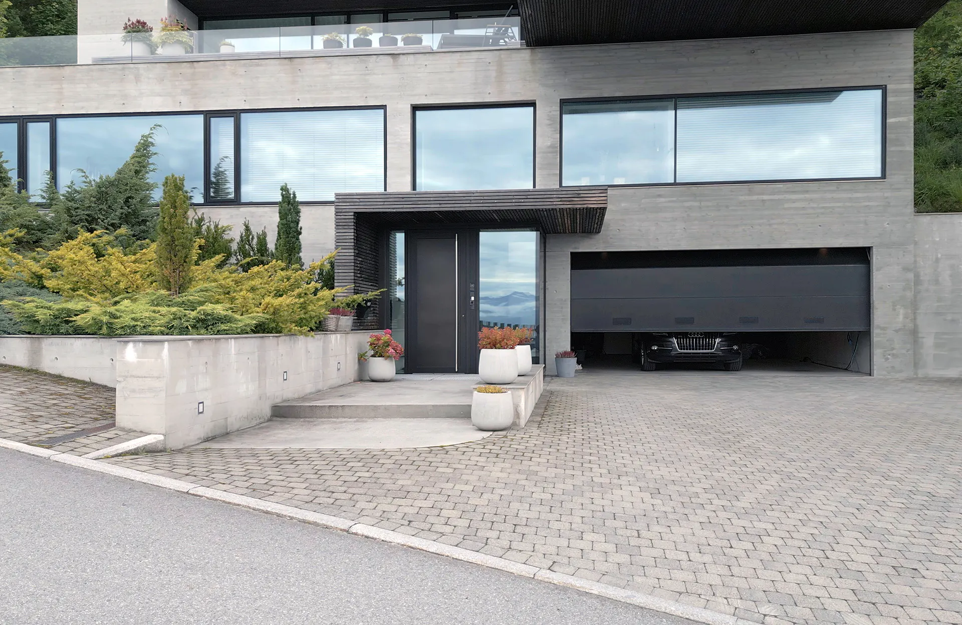 Store rektangulære vinduer med horisontale persienner dekker den øvre fasaden; en bred garasjeport er halvåpen og inngangspartiet med dør og overbygg ligger midt på en moderne betongvilla omgitt av prydbusker.