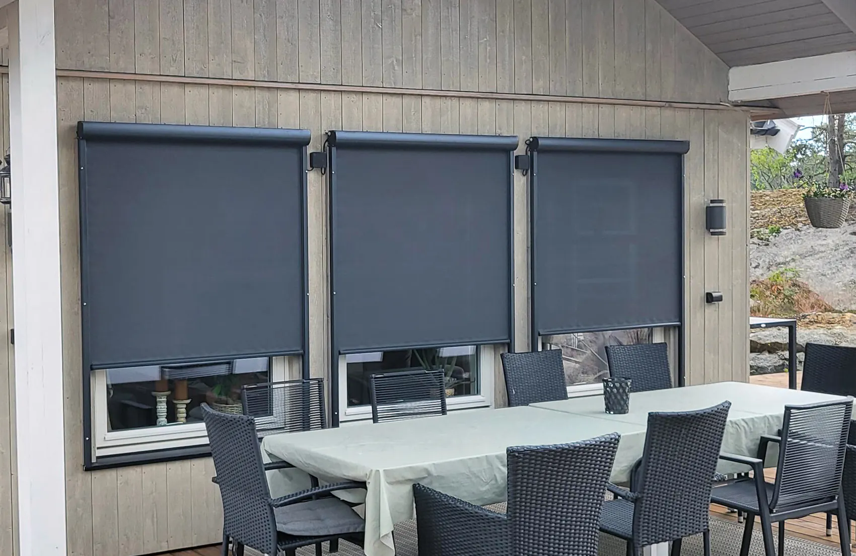 Tre mørkegrå utvendige screen er trukket ned over tre vinduer på en lys trefasade; foran står et dekket utebord med seks fletthavestoler på en terrasse.