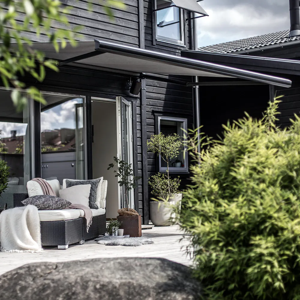 Kassettmarkise strukket ut over terrasse, skygger en koselig uteplass med kurvmøbel, puter og pledd. Mørkt trehus i bakgrunnen, åpen glassdør, potteplanter og stein i forgrunnen.