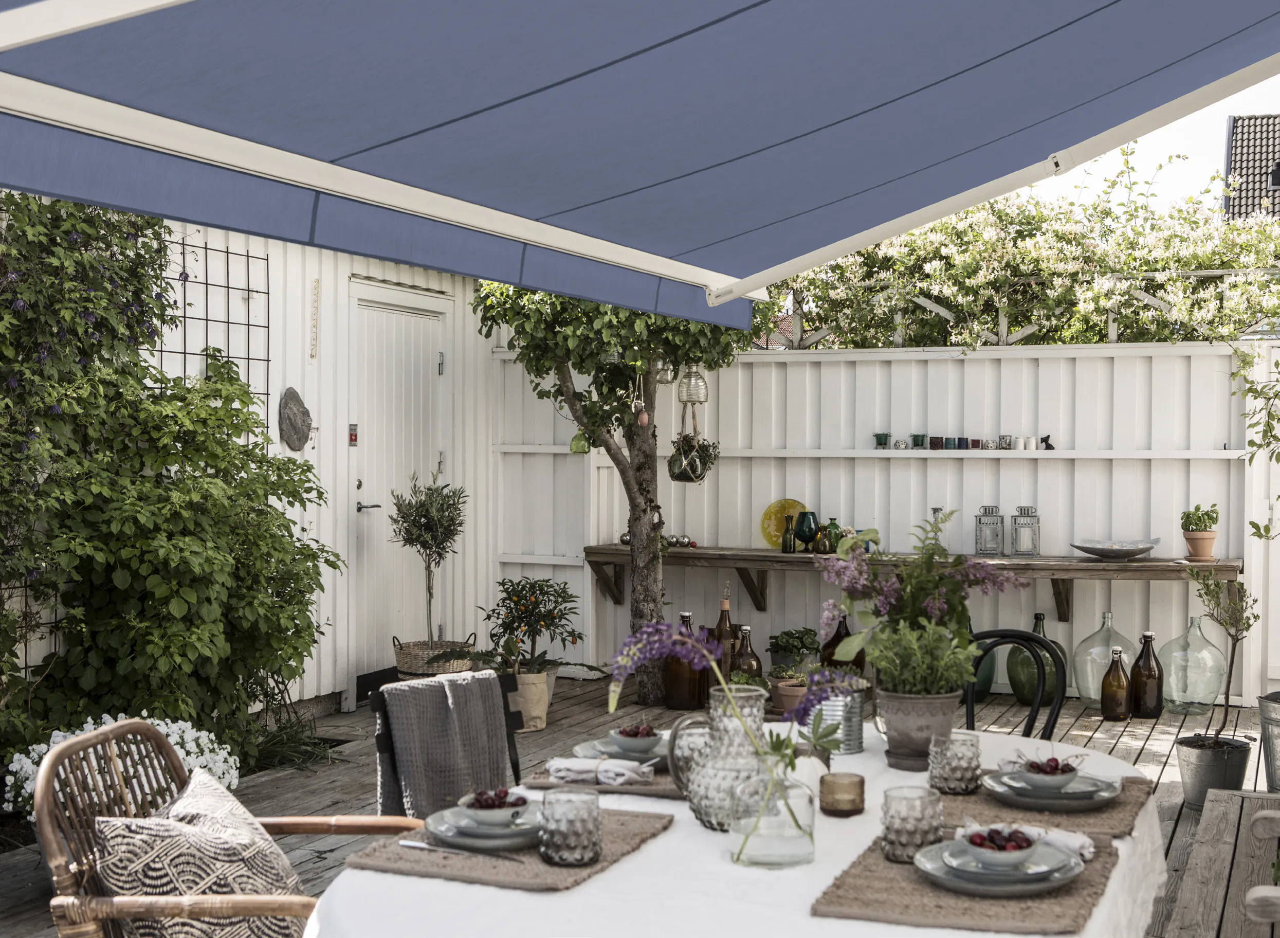 Markise i blått skygger over et dekket utebord. Terrasse med hvitt gjerde, hagemøbler, planter og dekorative krukker skaper en hyggelig, beskyttet uteplass.