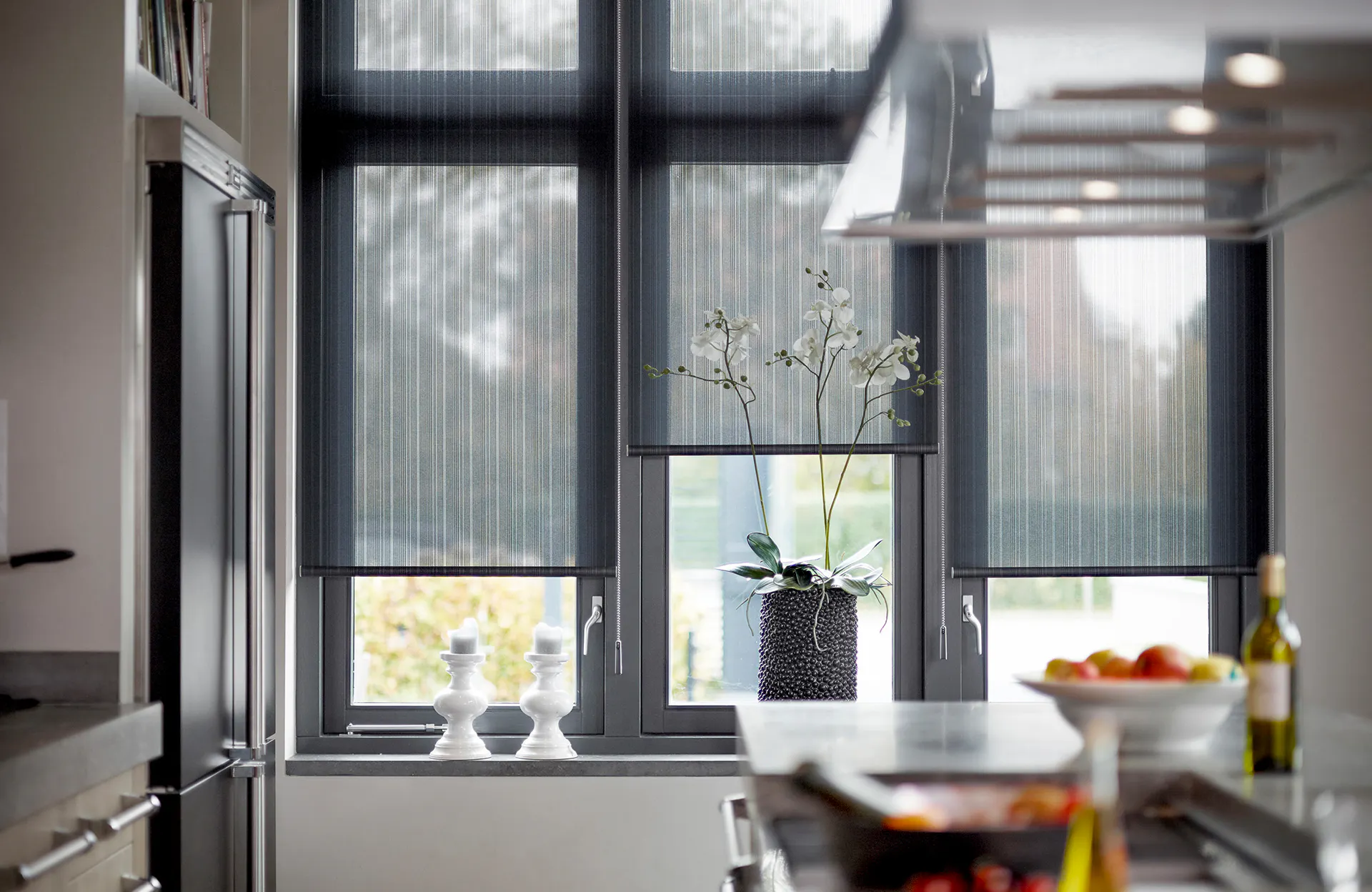Et mørkt, halvtransparent rullegardin dekker tre høye vinduer og filtrerer dagslys. I vinduskarmen står en mørk strukturert vase med hvite orkidéer og to lysestaker; kjøkkenbenk og pendellampe i forgrunnen.