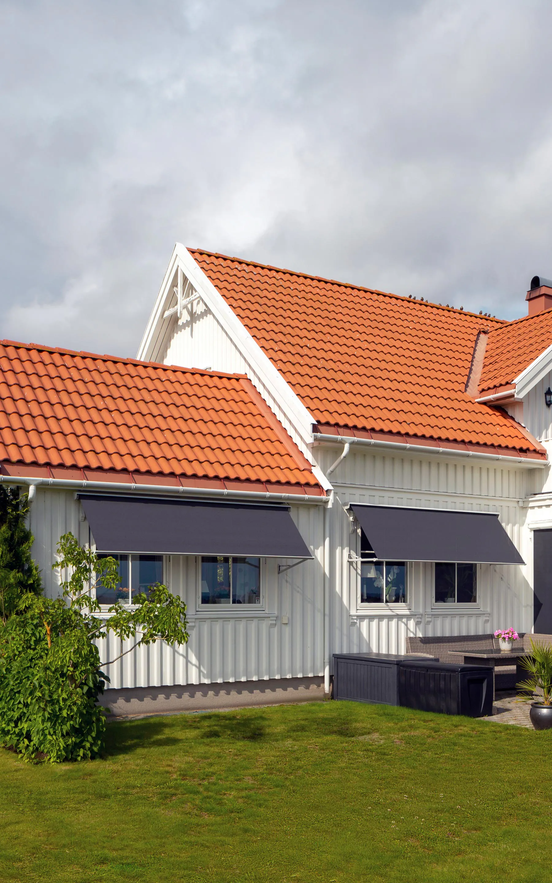 Markise trukket ut over balkongvindu på en hvit enebolig med oransje takstein; montert over inngangsparti, omgitt av gressplen og åpen, litt skyet himmel.