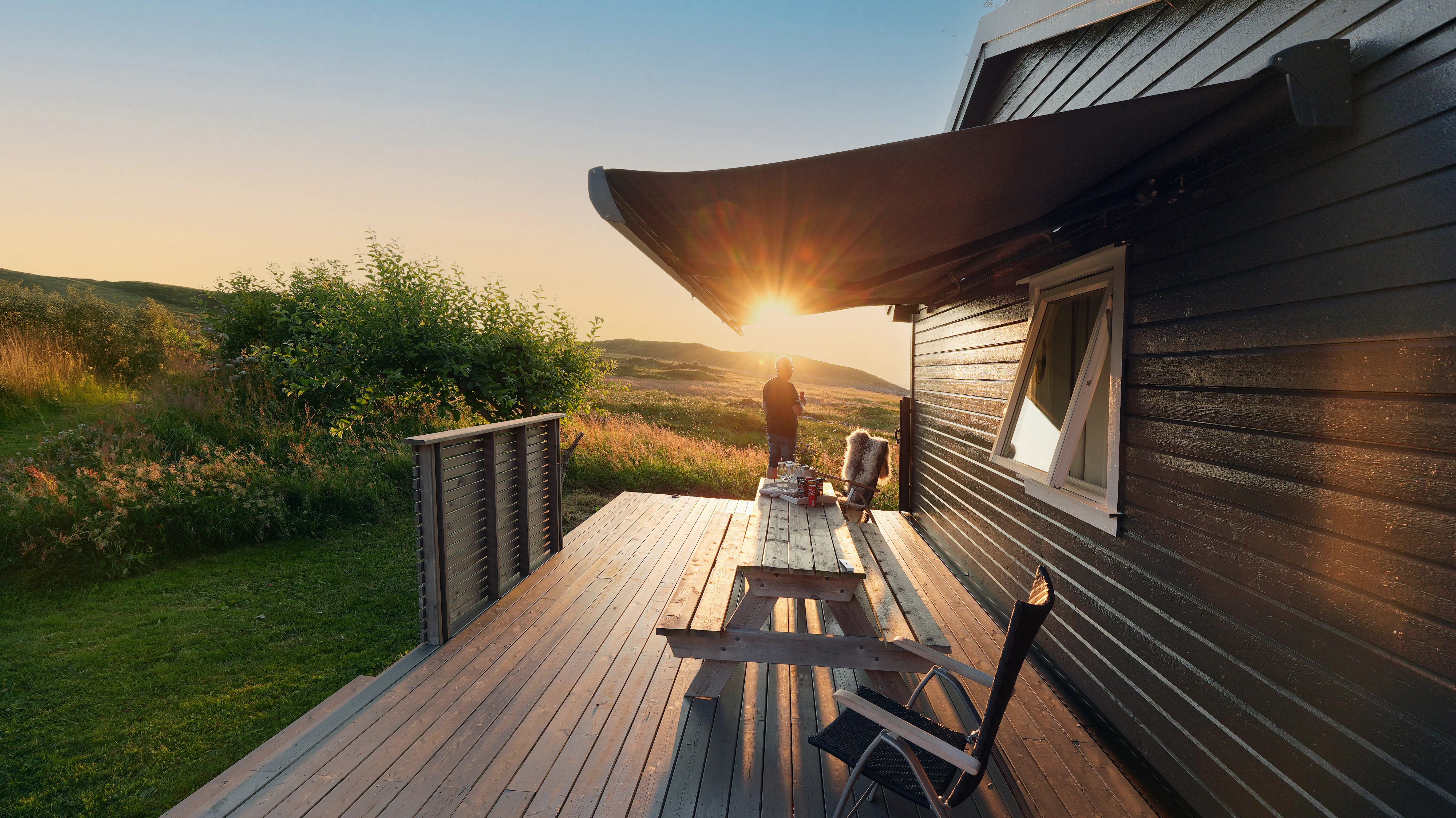 Markise er utstrakt over terrasse og gir skygge. Under står et trebenkbord og stoler; en person ved rekkverket ser mot solnedgang over gressdekte åser ved en husside i trepanel.