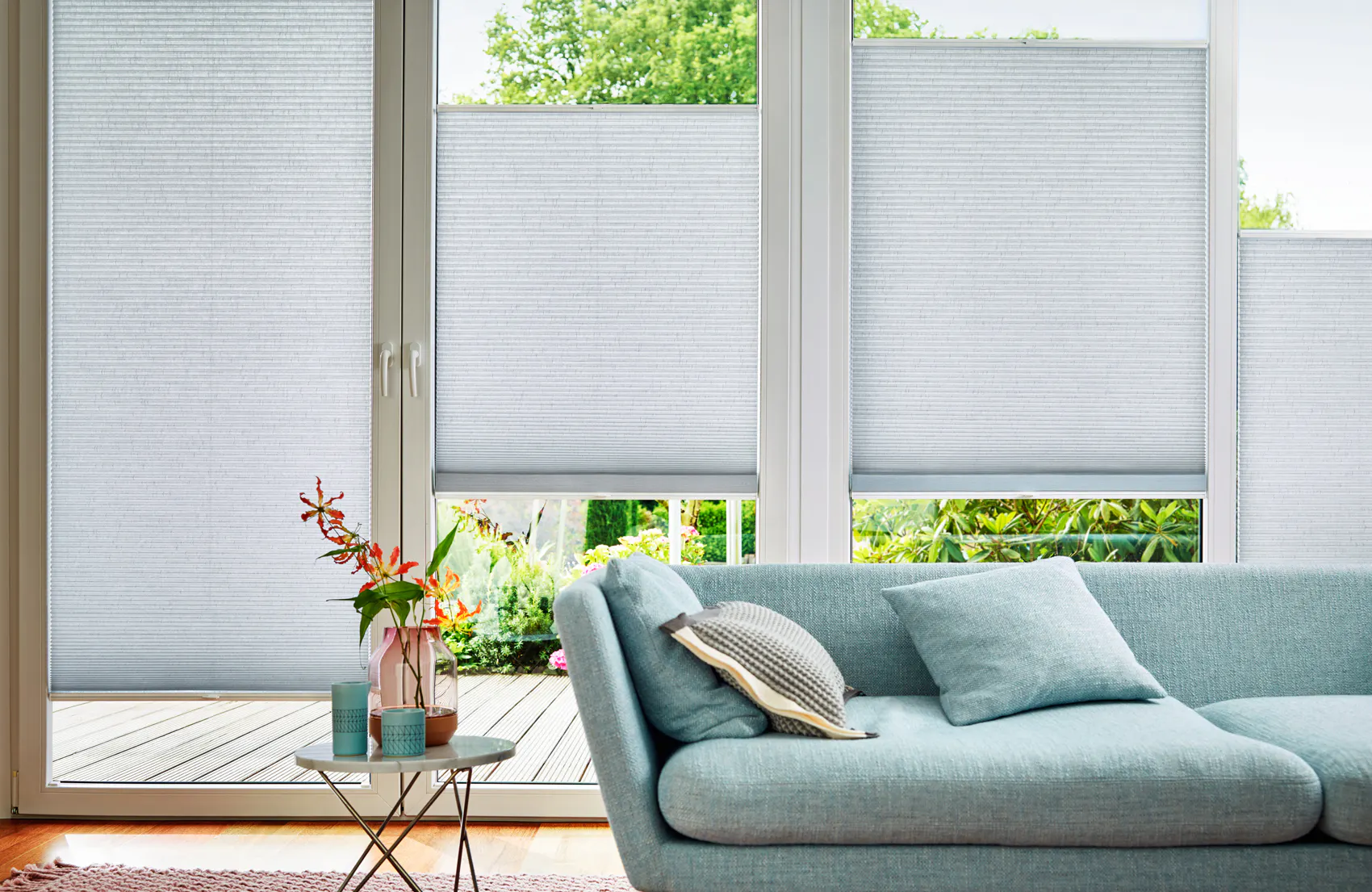 Plisségardiner dekker store franske vinduer og filtrerer dagslys, foran en lysblå sofa med puter; lite sidebord med vaser og utsikt mot en treterrasse og frodig hage.