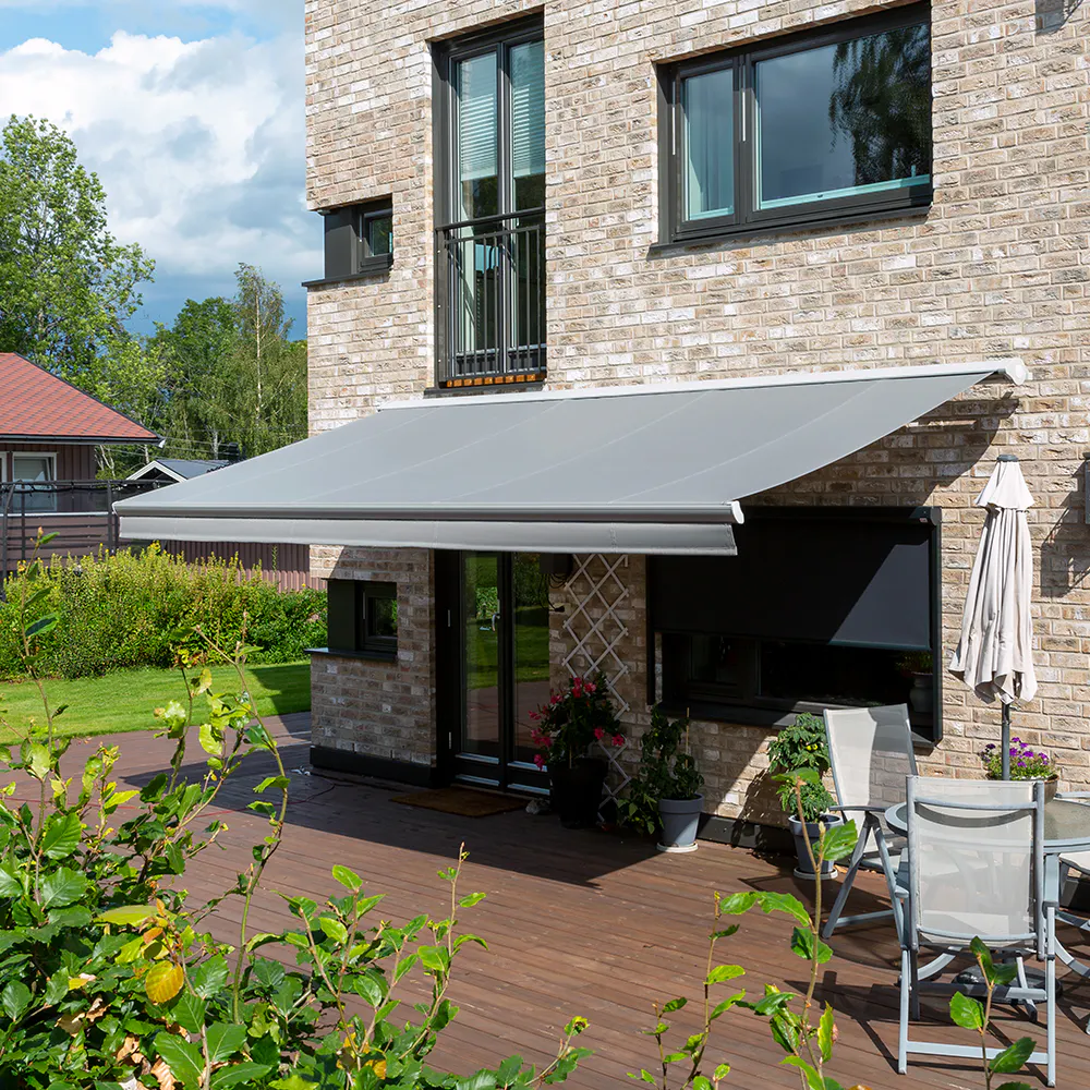 Terrassemarkise i grått utfelt over spisegruppe på tremurhus, med sammenslått parasoll ved siden