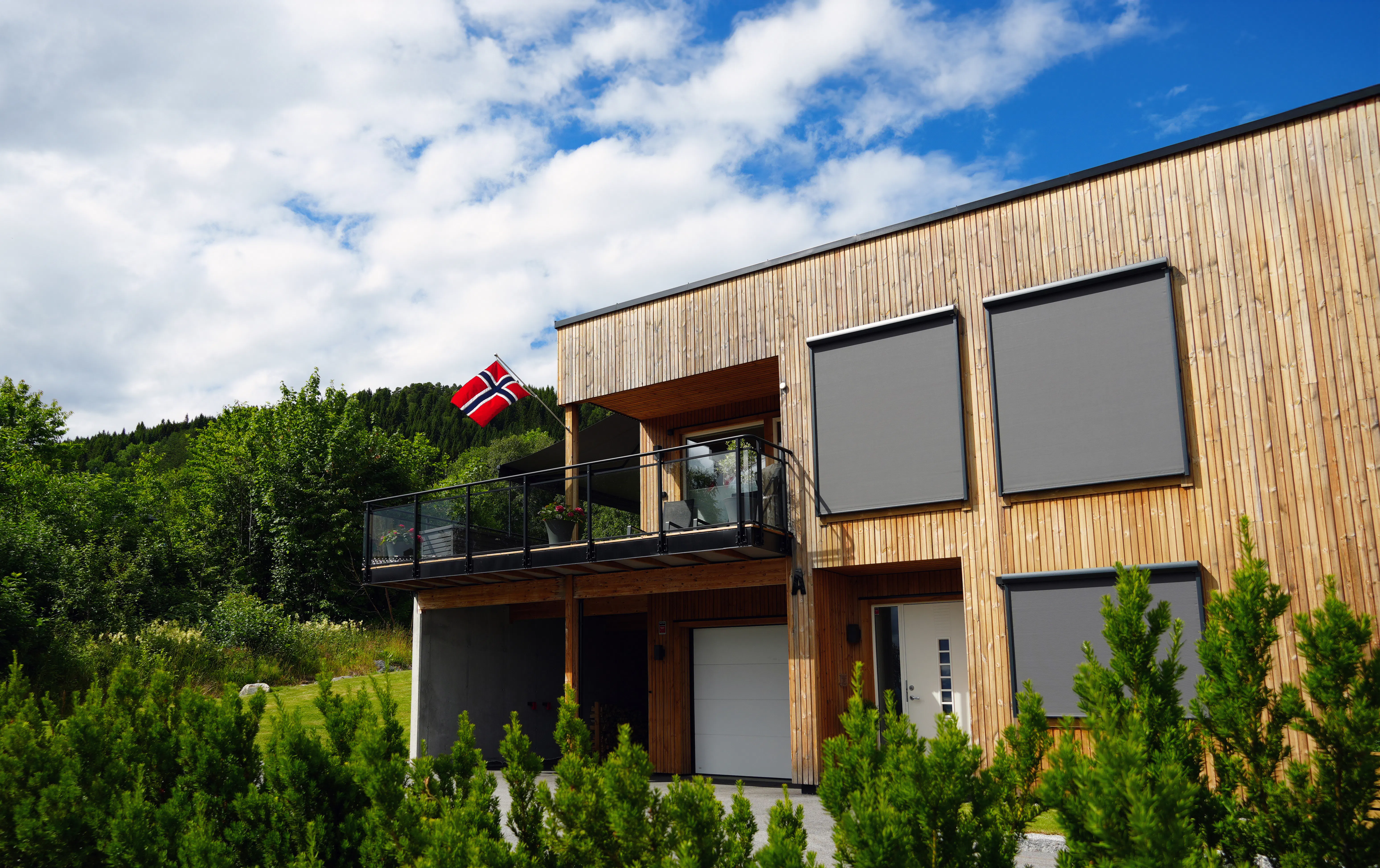 Utvendige screens er trukket ned foran vinduer på et moderne trekledd hus. Husets balkong har et norsk flagg; omgivelser med grønn beplantning, trær og blå himmel.