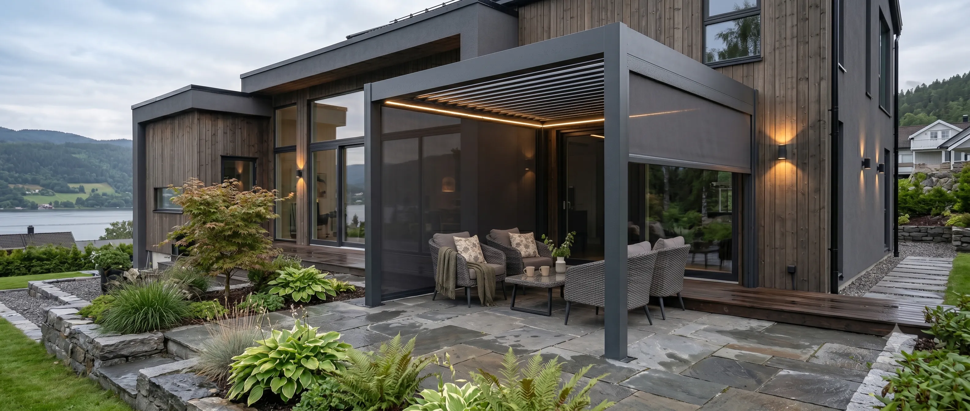 En moderne aluminiumspergola med nedrullede screens og integrert LED-lys dekker en sittegruppe med puter; står på skiferlagt terrasse foran en trefasade, omgitt av hageplanter.
