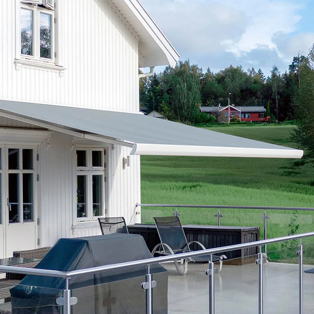 Uttrekkbar hvit markise strekker seg ut over en terrasse og skygger hagemøbler. Hvitt trehus med vinduer og glassrekkverk mot en grønn eng og et rødt hus i bakgrunnen.