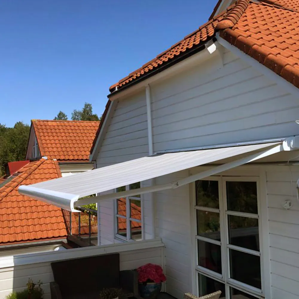 Hvitt kassettmarkise rullet ut over balkong, gir skygge til møbler og potteplanter foran hvit trefasade med franske dører; rød takstein og blå himmel i bakgrunnen.