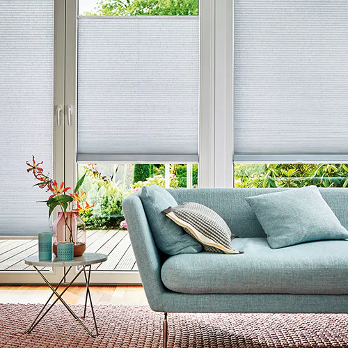 Duettegardiner er trukket ned over store glassdører og blokkerer mesteparten av lyset fra en terrasse. Rommet har en lys blå sofa med puter og et lite sidebord med blomster.