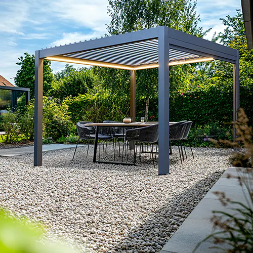 En grå pergola med lamelltak gir skygge og varmhvitt LED-lys over et spisebord med seks stoler på en grusdekket uteplass omgitt av hekk og hage.