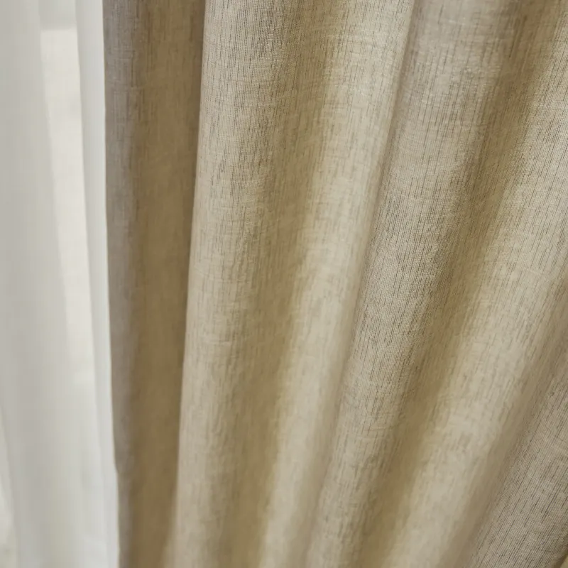 Beige tekstilgardin henger i myke, vertikale folder foran en hvit, gjennomsiktig voilage; nærbilde viser stoffets fine struktur og svakt filtrert dagslys i et innendørs vindusmiljø.