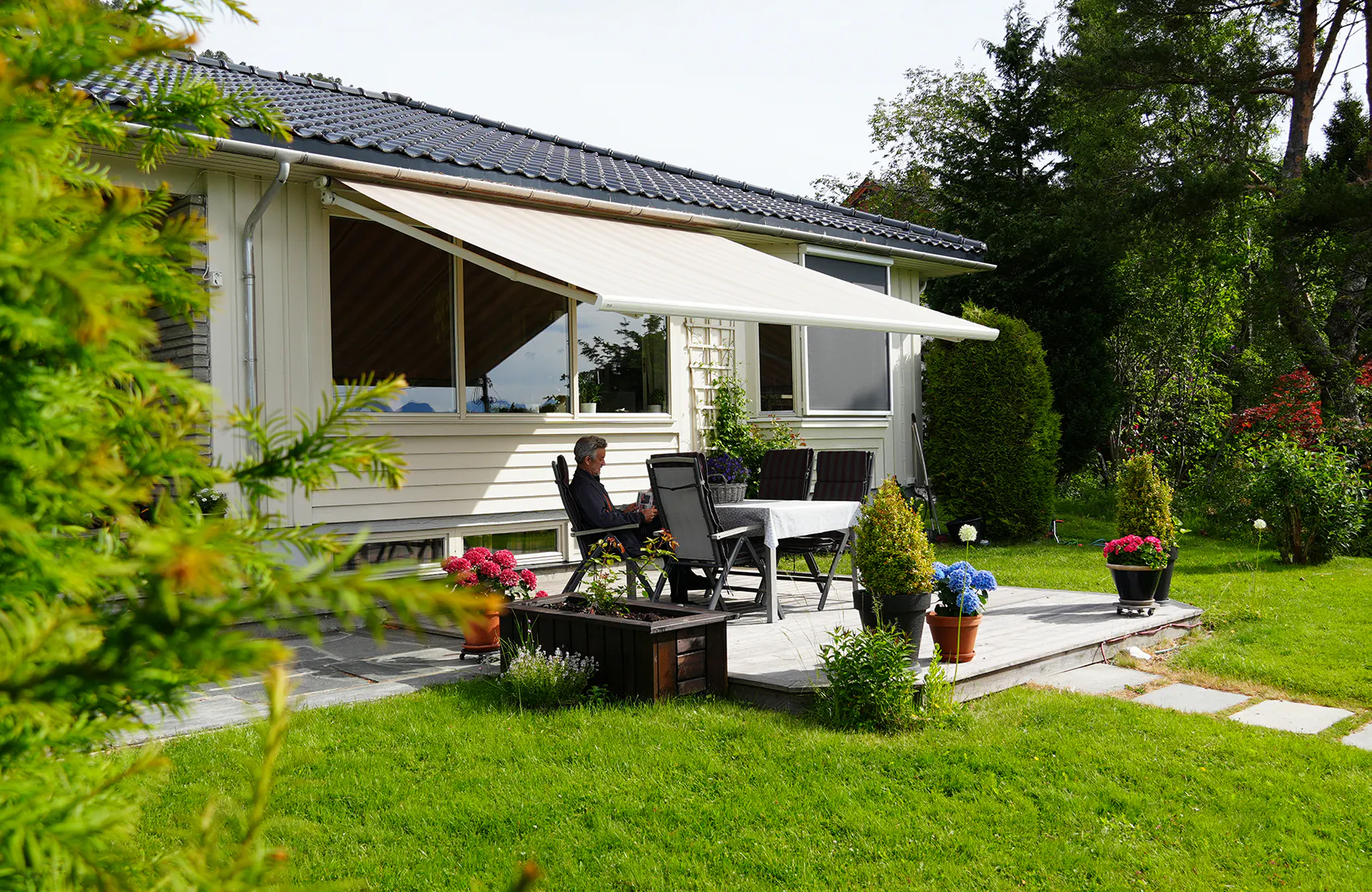 Markise gir skygge over terrasse; en person sitter ved hagebord med stoler. Enetasjes hvitt trehus flankert av grønn plen, potteplanter og trær i en rolig hage.