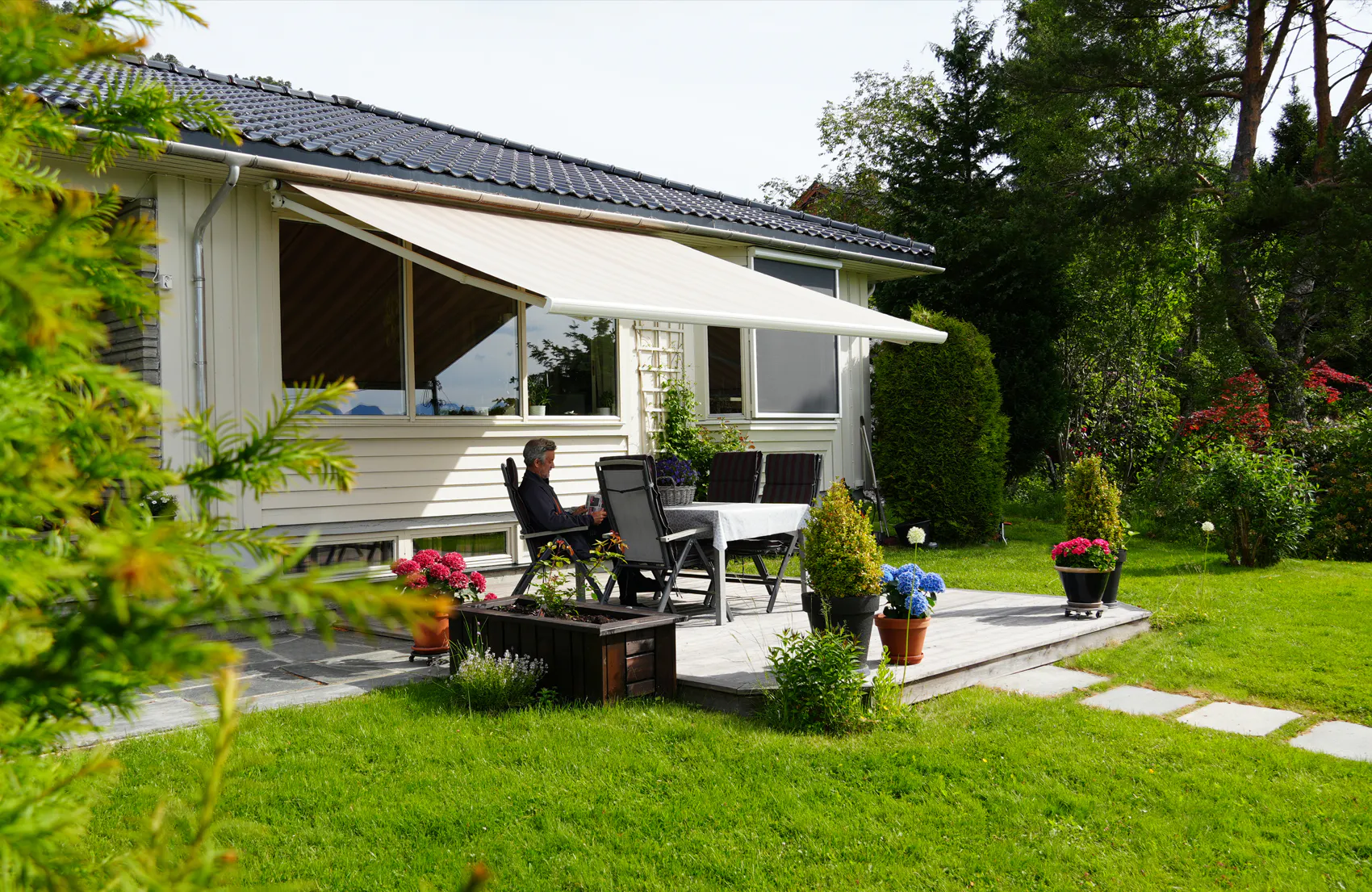 En hvit terrassemarkise er utfelt og skygger en uteplass; et bord med stoler og en person sitter ved bordet. Hvitt enetasjes hus med store vinduer, grønt gress og blomsterkrukker.