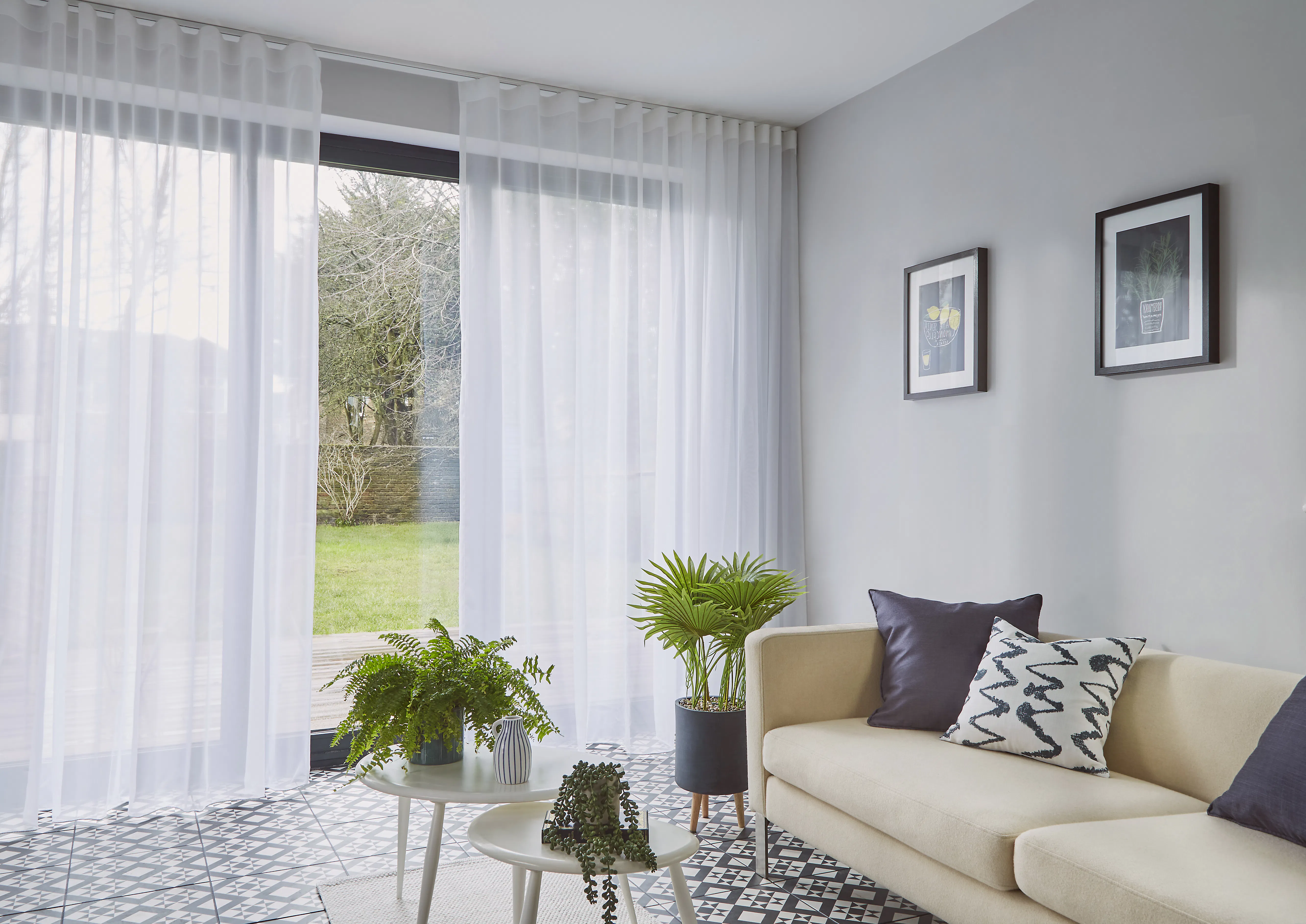 Lette, hvite gardiner henger foran en skyvedør og demper dagslys; stue med lys sofa, mønstrete gulvfliser, grønne potteplanter og to rammede bilder på grå vegg.
