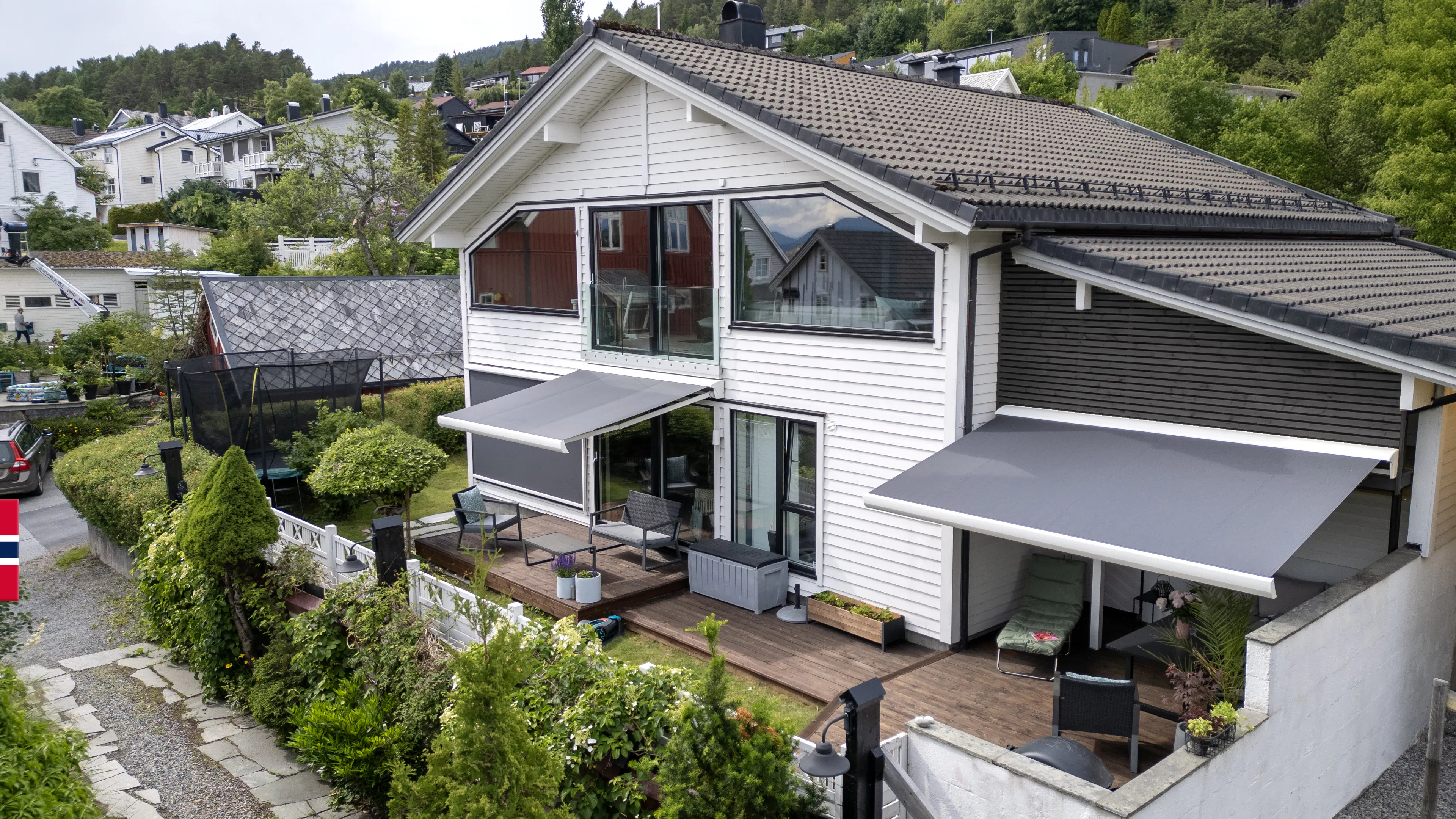 To grå markiser dekker terrasse og balkong på hvit enebolig, skaper skygge over utemøbler på tredekke. Hus ligger i grønt forstadsområde i skrånende terreng med nærliggende hus.