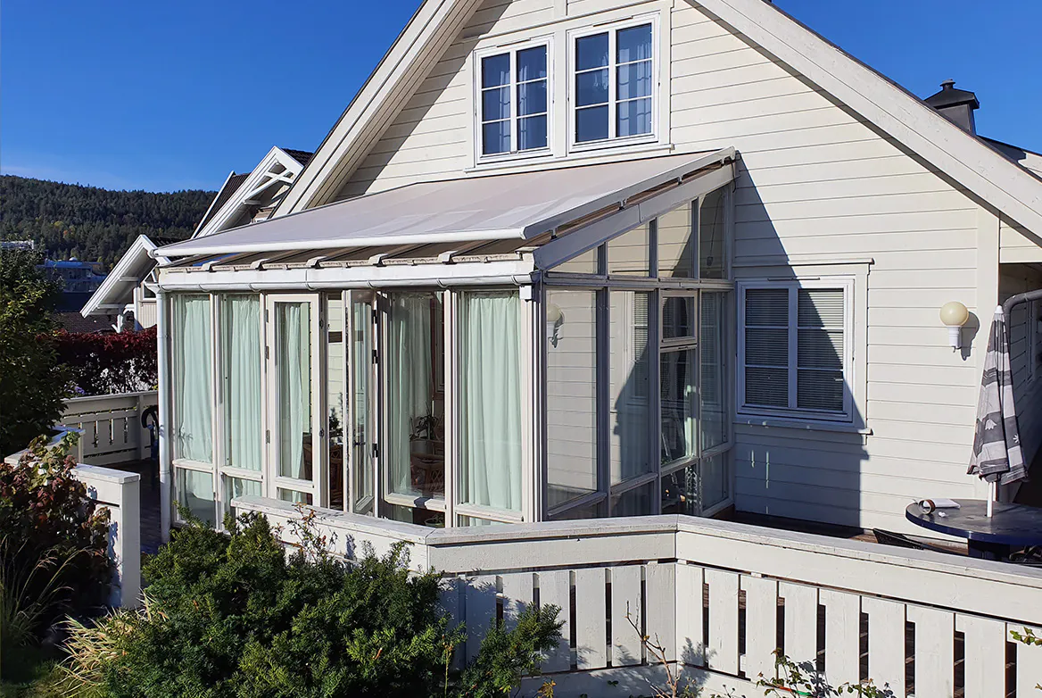 Vertikale lamellgardiner dekker glassvegger i en innglasset veranda festet til et hvitt trehus; verandaen vender mot en terrasse med hvitt gjerde, busker og klar, blå himmel.