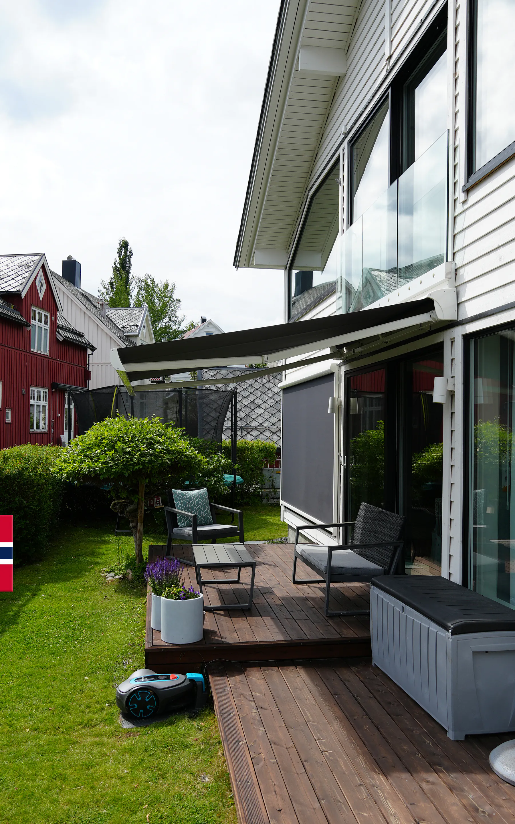 Markise med nedrullet screens montert over en terrasse; markisen er utfelt og gir skygge, screens beskytter mot innsyn. Moderne hvit bolig med glassdører, terrassemøbler, gressplen og nabohus.