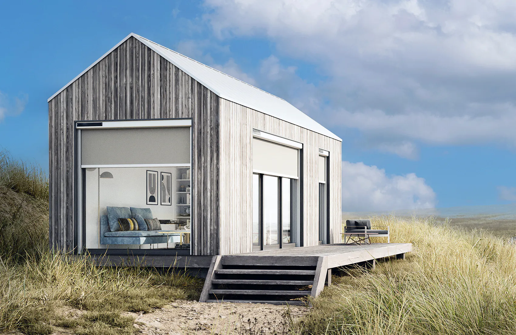 Utvendige rullgardiner på en moderne, gråtrekledd hytte; gardinene er delvis senket foran store vinduer. Hytta står på en treplattform ved sanddyner og gress under blå himmel.