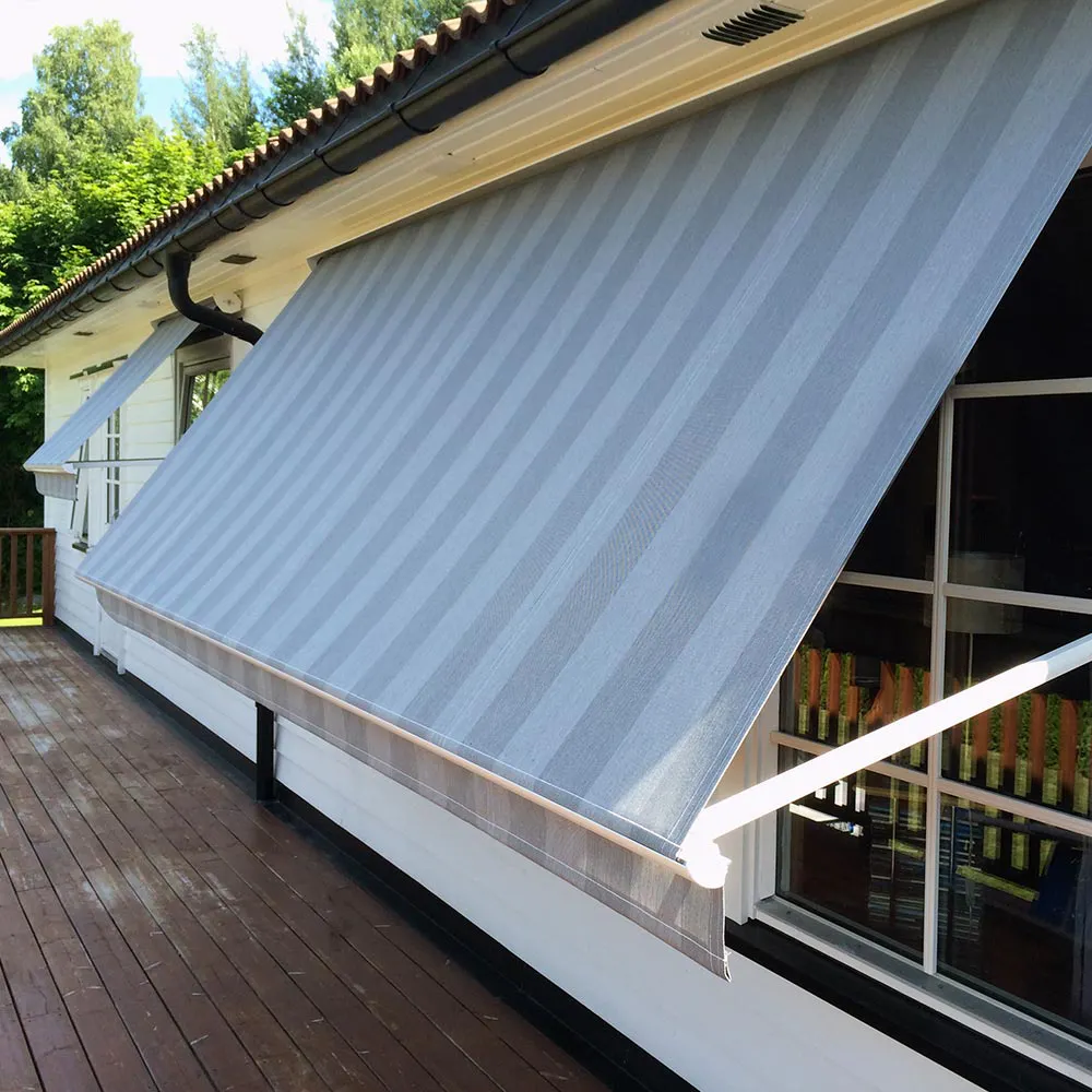 Utvendig, stripete lys grå/blå markise strukket ut over fasadevindu, gir skygge til vinduer og treterrasse. Montert under takrenne på hvitt hus med grønn vegetasjon i bakgrunnen.