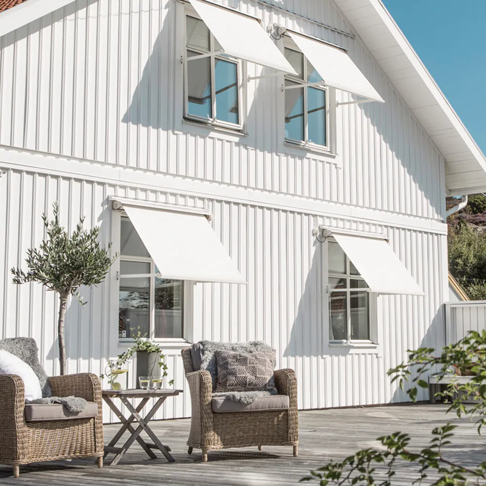 Vegghengte hvite markiser dekker fire vinduer, trukket ut for å gi skygge. Hvitt, liggende panelt trehus med solrik terrasse, flette lenestoler, lite bord og potteplanter.
