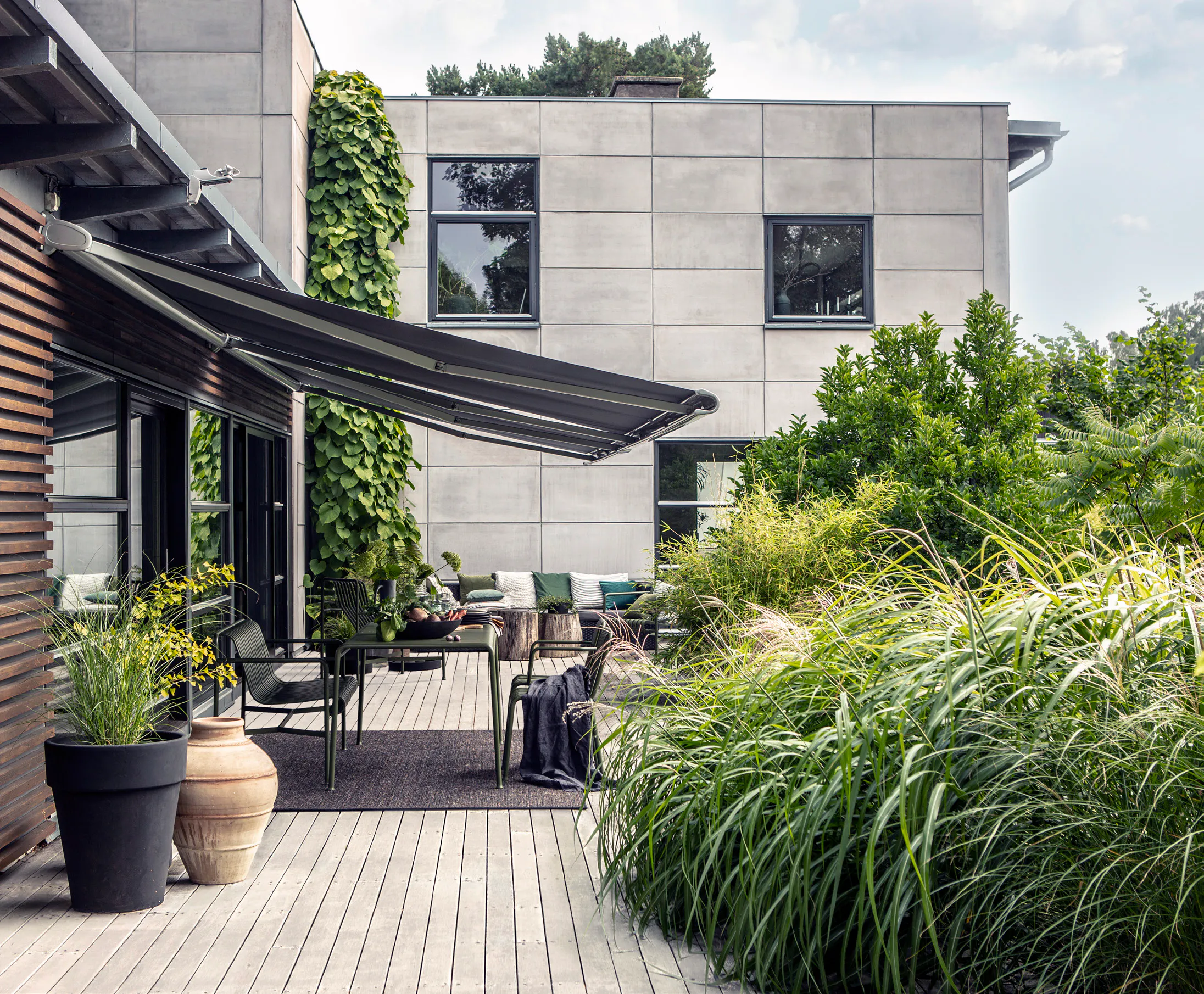 En uttrukket terrassemarkise skygger et uteområde med bord, stoler og puter; treplatting og store krukker ved en moderne grå fasade med vinduer og frodig grønn beplantning.