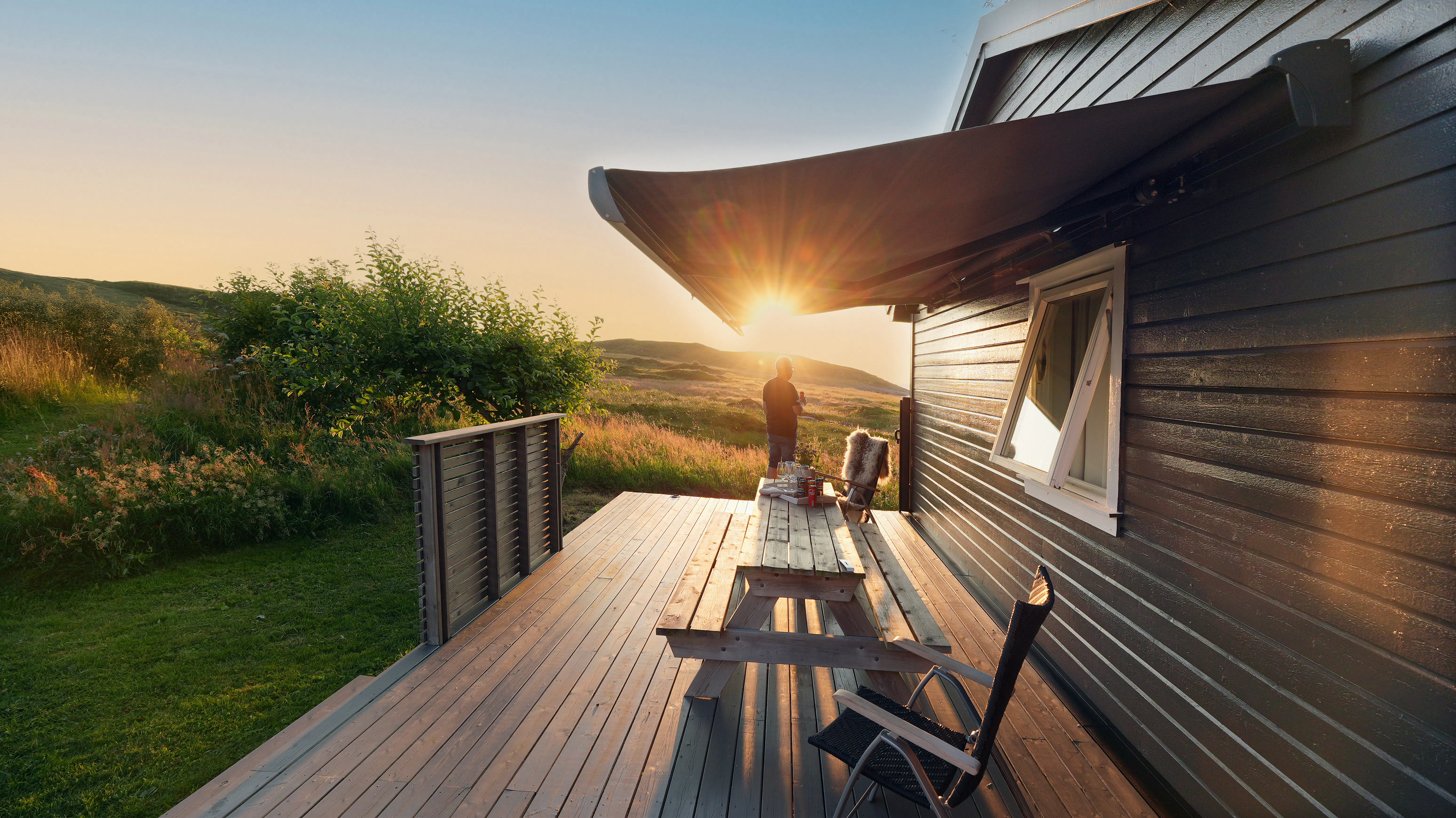 Markise - skygger en treterrasse ved et mørkt, trekledd hus; piknikbord og stol står under, mens solen synker bak gresskledde åser og åpne landskap i bakgrunnen.