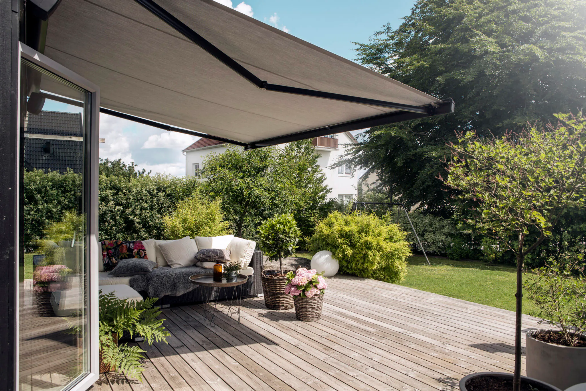 Kassettmarkise er uttrukket over en treterrasse og skygger en uteplass med hjørnesofa, bord og blomsterkurver; omgir av grønt buskas og plen ved en enebolig på en solrik dag.