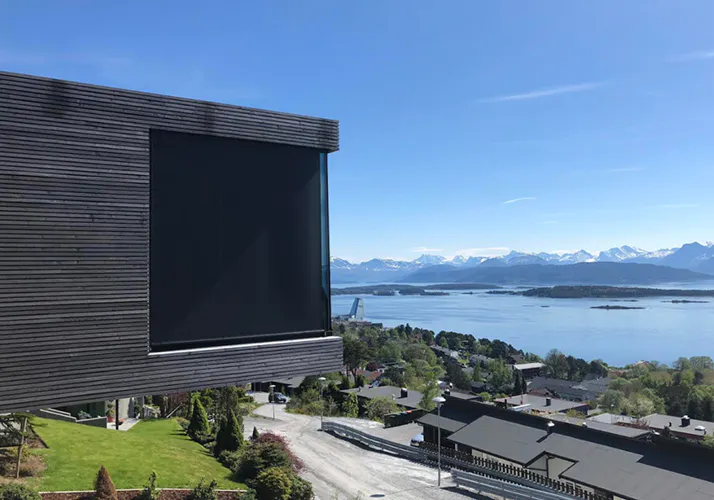 Utvendig rullegardin senket over et stort vindu i moderne trekledning; i kontekst en åpen kystlinje med boliger, vei, sjø, øyer og snødekte fjell under klar blå himmel.