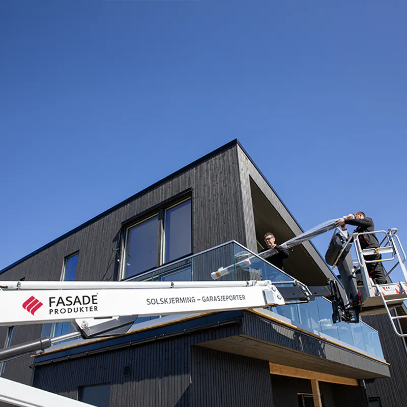 Glassrekkverk monteres (handling) av arbeidere i lift (handling) foran et moderne, mørkt trekledd hus under klar blå himmel (kontekst).