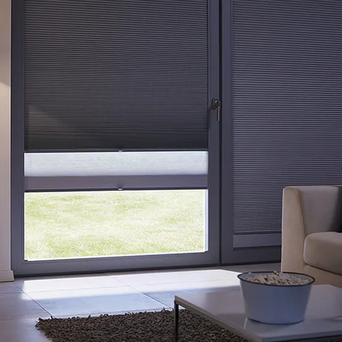 Mørk plisségardin delvis senket på en glassdør, med en lys gjennomsiktig stripe som slipper inn dagslys og viser gress utenfor; stueinteriør med sofa, teppe og sofabord.