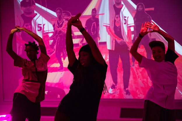 Three performers dance in unison with their right hips jutted out and their hands held together above their heads. The two performers on the outer edges wear white t-shirts and the performer in the center wears a black t-shirt. Behind them, a projection against the wall protrays five other people walking. The entire scene is lit with pink light. 