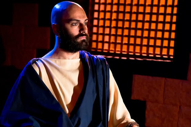 Portraiture shot of a bearded person with a shaved head dressed in biblical garb, facing ahead in a dim room with a persimmon orange wall.