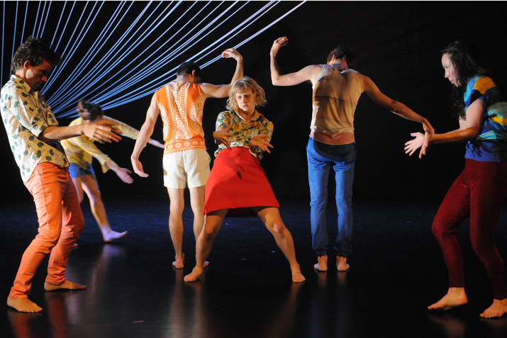 A performer in a red skirt stands in the center of a dimly lit stage surrounded by dancers posing symmetrically. On the performer's side, two performers face each other fanning out their palms. Behind the performer, two performers face backwards each raising one bent arm and holding out the other. 