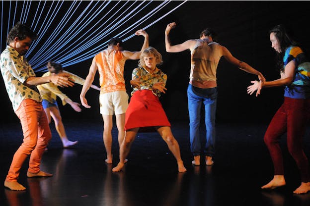 A performer in a red skirt stands in the center of a dimly lit stage surrounded by dancers posing symmetrically. On the performer's side, two performers face each other fanning out their palms. Behind the performer, two performers face backwards each raising one bent arm and holding out the other. 