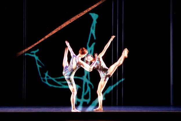 Two performers are in the center a dark stage wearing leotards with a metallic blue toned abstract pattern on them. Looking directly at each other, they lean to the side on one leg while lifting their other leg. They are mirroed. Behind them, a blue sketch of a human figure is projected onto the otherwise black wall. This figure is upside down and only its legs are visible. 