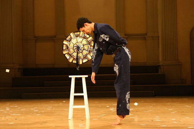 Jonah Bokaer performs, wearing a blue jumpsuit stained with white paint footprints. He stands on his tip toes and bends over, extending his right hand downwards. Behind him, there is a wheel of metrocards on top of a white stool. The wood paneled floor beneath him is littered with metrocards. 