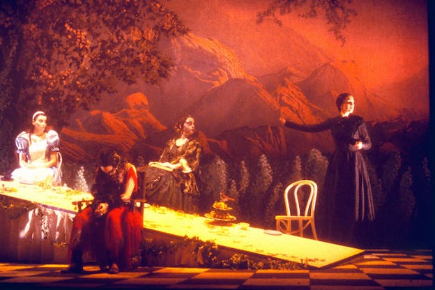 Four people stand in front of a table that has collapsed on its right end. One woman dressed in red has her back turned towards the rest and is looking at the floor. Another one clad in black has stood from her chair while the other two look in her direction. There are mountains in the background and everything is washed with a red hue.