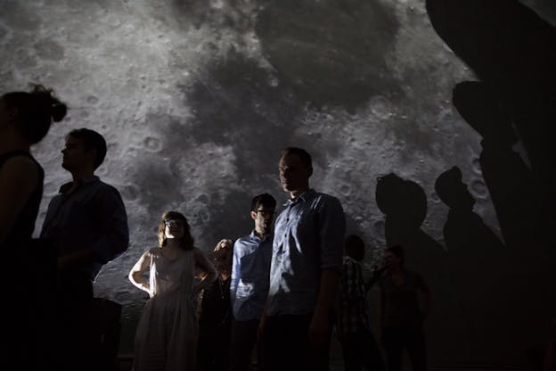 Three figures stand in a dark space with a projection of the moon behind them. Shadows of other figures obscure parts of the projection.