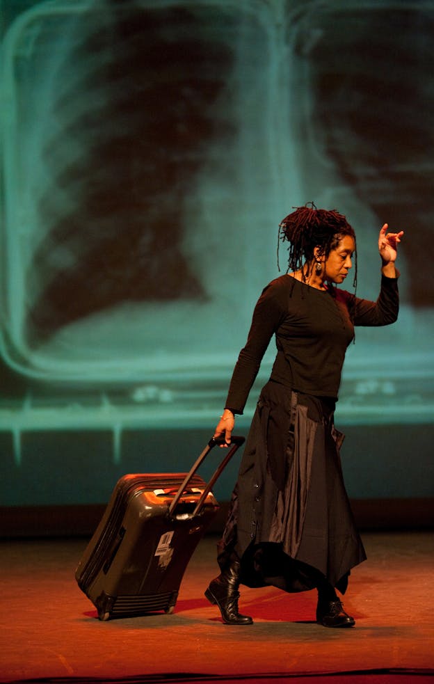 A photograph of Pamela Z performing, wearing a black long sleeved shirt and a draped black skirt and draging a black suitcase behind her. She appears mid-stride and has one arm raised by her head. Behind her, an x-ray of a rib cage is projected on the wall. The floor is illuminated with orange-red light. 