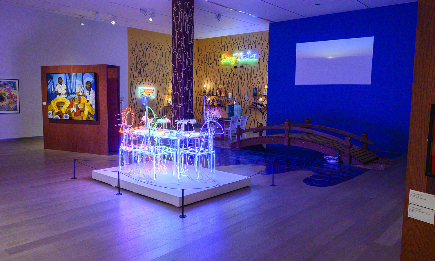A neon table is lit up in the center of the room, illuminating a blue wall with a projection of a sunset and a bridge built directly onto the floor. The bridge leads to the next room, which has neon signs and various objects sitting on shelves affixed to the wall.