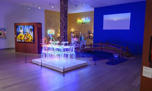 A neon table is lit up in the center of the room, illuminating a blue wall with a projection of a sunset and a bridge built directly onto the floor. The bridge leads to the next room, which has neon signs and various objects sitting on shelves affixed to the wall.