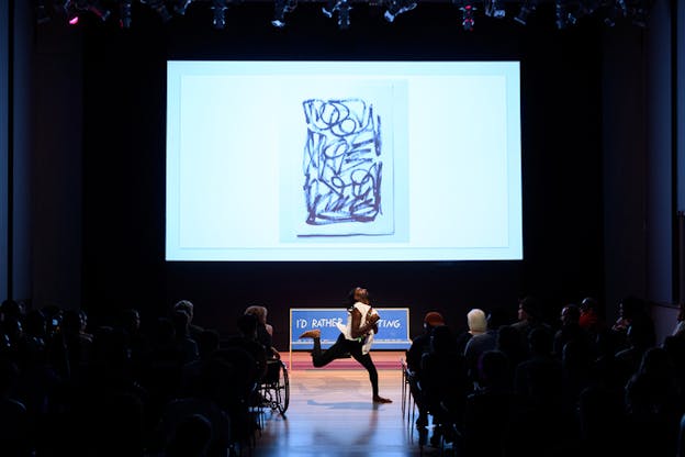 A performer moves in the middle of the stage, the audience sitted and separated in two groups on each side giving a clear view of them. Behind them a projection of abstract art, a black swirled conitnuous line on a white canvas.