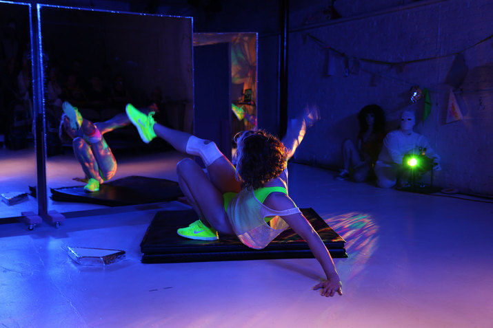 Person dancing before a mirror in a darkened, purple-hued room. In the background a neon green light illuminates the dancer's neon green attire and casts colorful strobes of light on the dancer's body and on the floor.