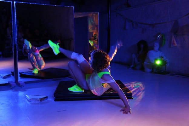 Person dancing before a mirror in a darkened, purple-hued room. In the background a neon green light illuminates the dancer's neon green attire and casts colorful strobes of light on the dancer's body and on the floor.